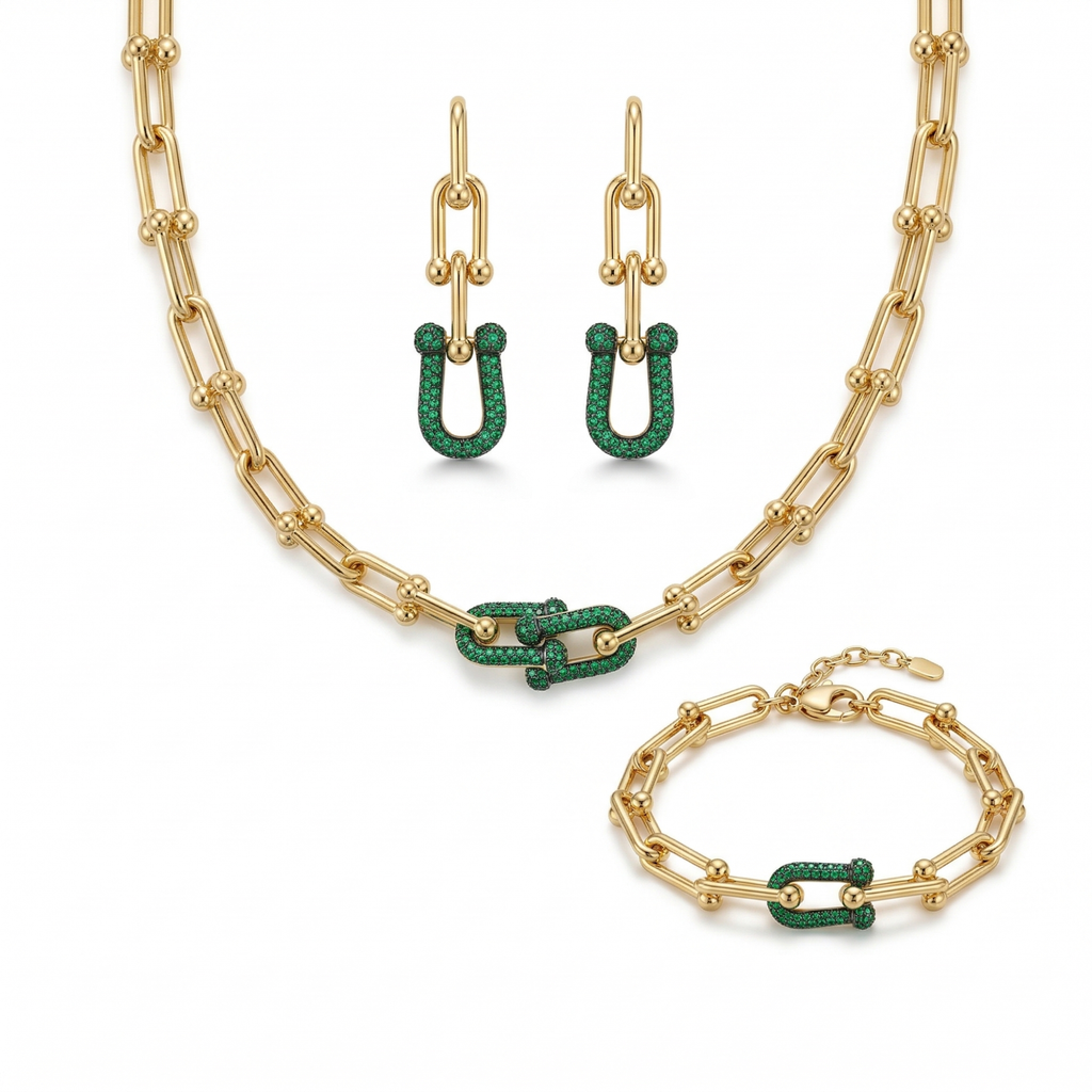 Un conjunto de joyas de lujo de acero bañado en oro de 18K, compuesto por un collar de cadena de eslabones gruesos, una pulsera a juego y un par de pendientes largos. Cada pieza tiene un diseño central de eslabones entrelazados adornados con pavé de piedras verdes brillantes, fabricado en Italia.